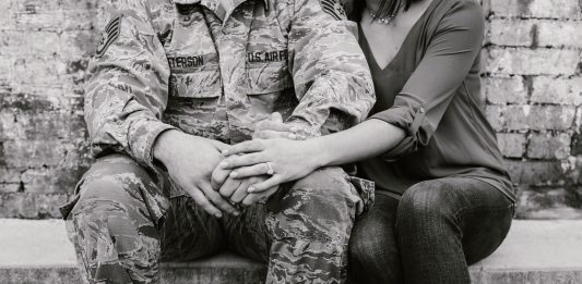 Military spouse mother working remotely on a laptop while her child plays, demonstrating a balanced home and career life.